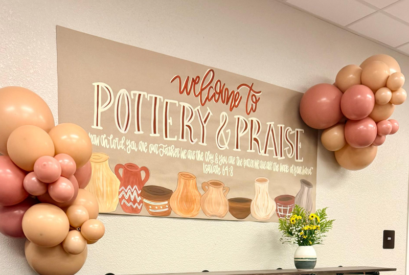 Decorative setup with balloons and a table in a room with a 'Welcome to Pottery & Praise' sign.
